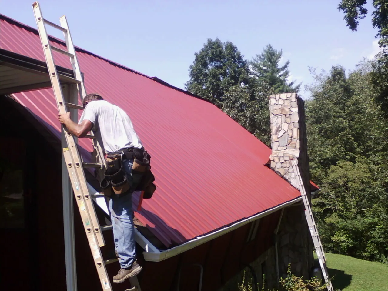 Roofer on a ladder beside a red metal roof and stone chimney — Soffit & Fascia Repair by Roofing Brothers in New Lisbon
